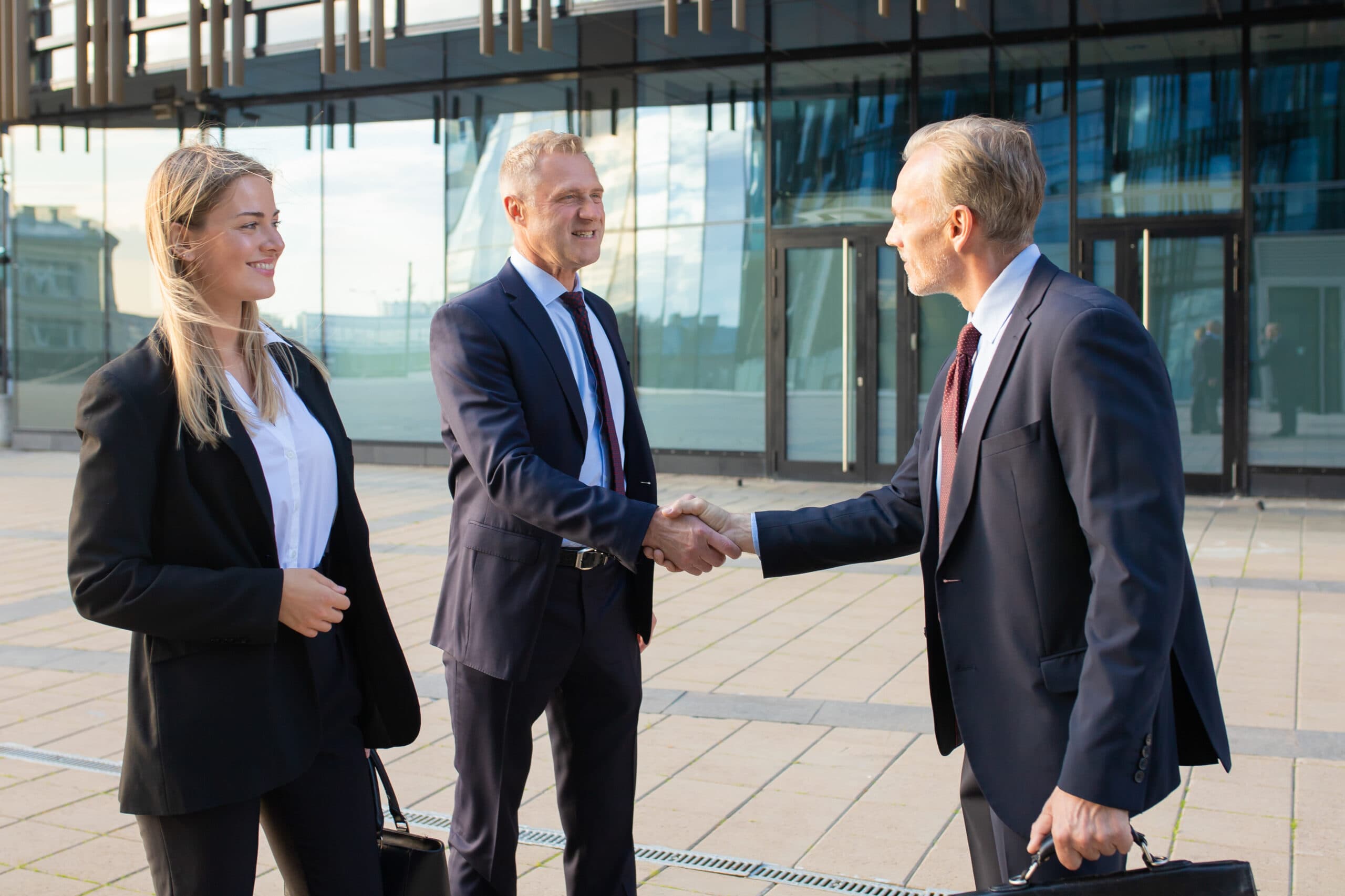 A female professional standing with two male professionals who are shaking hands outside a commercial office building.