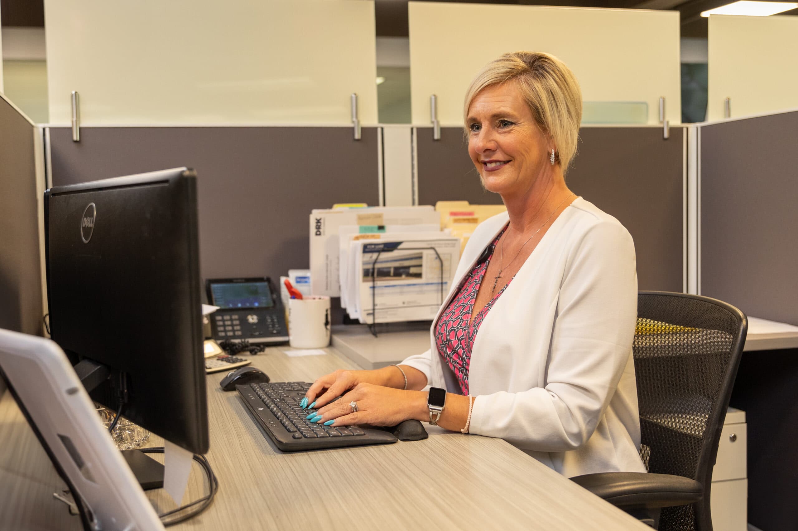 image of a woman with short blonde hair sitting at a desk while typing on a keyboard and looking at her computer screen.