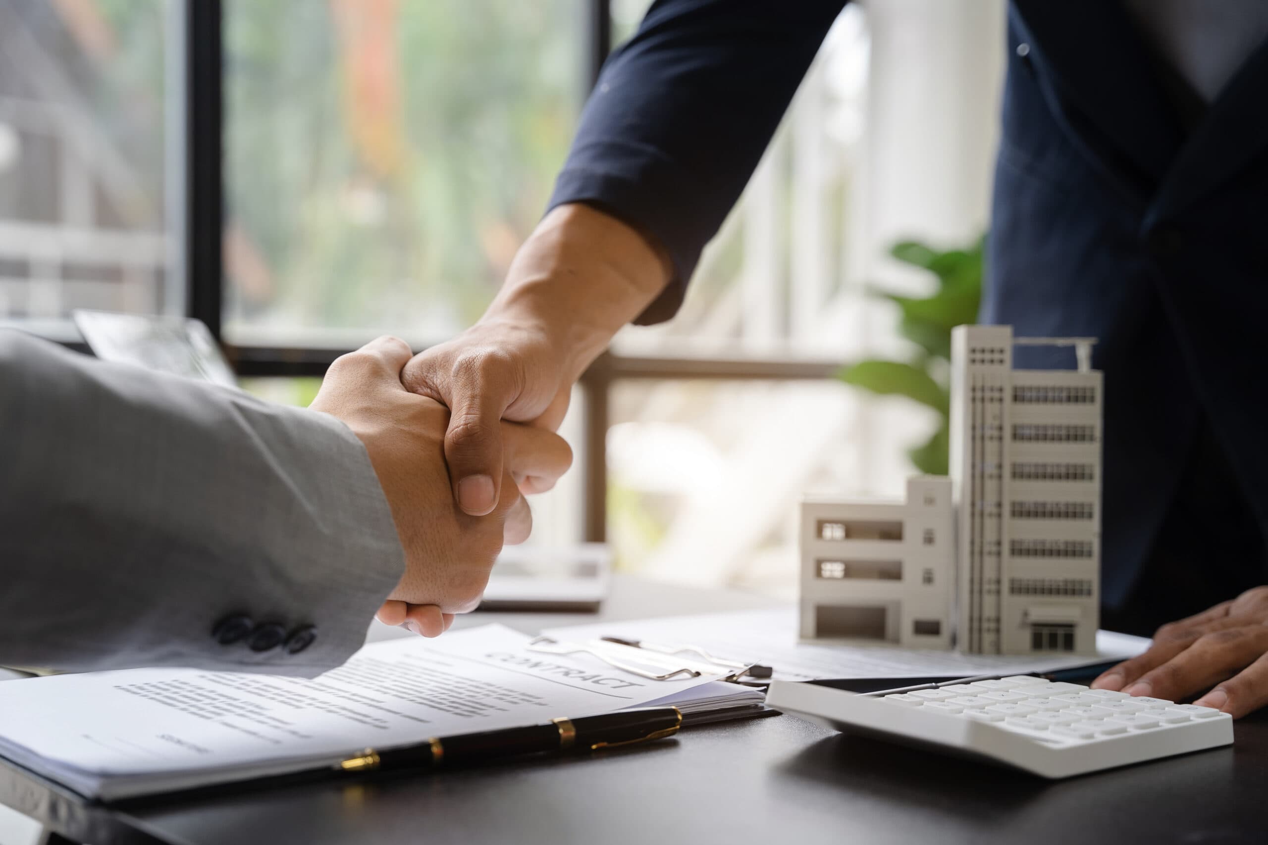 Two individuals shaking hands across a desk with paperwork, a calculator and a building model on the table.